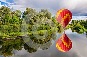 A bright hot air balloon flies low over the river black grouse throwing its reflection into  water