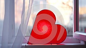Bright lamp in shape of heart is on window. Concept. Red heart-shaped lamp stands at window and glows to bestow love on