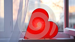 Bright lamp in shape of heart is on window. Concept. Red heart-shaped lamp stands at window and glows to bestow love on