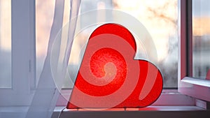 Bright lamp in shape of heart is on window. Concept. Red heart-shaped lamp stands at window and glows to bestow love on photo
