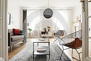 Bright living room interior with a metal armchair, coffee table and sofa. Big lamp in the middle. View through a d