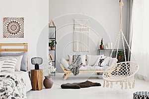 Bright living room with macrame on the wall, beige couch with pillow and blanket, hammock chair, fluffy rug and bedside