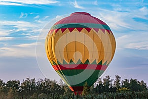 A bright multicolored hot air balloon at the moment of light touch with the ground on a forest lawn against a blue sky during suns