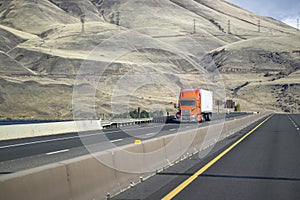 Bright orange bonnet big rig semi-truck with grille guard transporting cargo in dry van semi-trailer climbing uphill on the photo