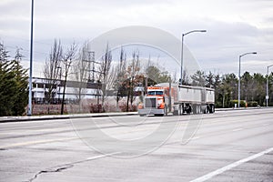 Bright orange classic American bonnet semi truck transporting two bulk semi trailer loaded with cjmmercial cargo driving on the