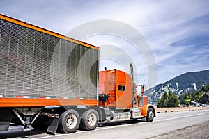 Bright orange classic American idol big rig semi truck with chrome parts transporting cargo in same color reefer semi trailer