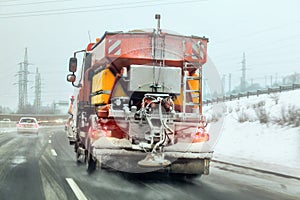 Bright orange gritter maintenance truck spreading deicing salt and sand on highway, view from car driving behind