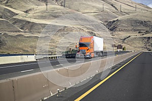 Bright orange long haul big rig semi truck with grille guard transporting cargo in dry van semi trailer running on the highway