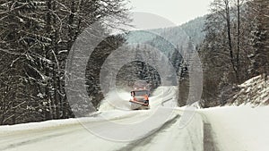 Bright orange plough truck cleaning snow on forest road in winter, view from car driving other way, trees around