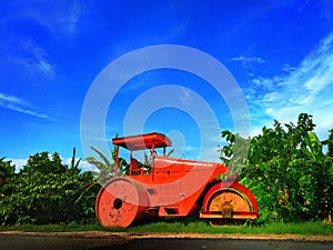 Bright orange road roller stands on not ready new road, stones, blue sky, cloud. Old wheels. India