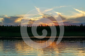 Bright orange sunset behind the clouds of sun on the banks of the Northern river Viluy under the blue sky in the spruce forest