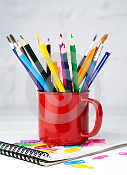 Bright pencils and brushes in a red mug on a white wooden table. School supplies. Back to school. Close-up. Selective focus