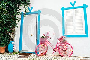 Bright pink bicycle decorated with artificial flowers standing near a white wall with closed window and door of