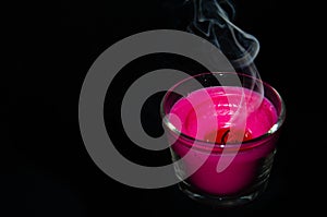 Bright pink candle in a glass jar on a black background. The candle is burning. Extinguished candle. Smoke from the candle. Hearth