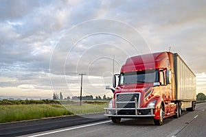 Bright red big rig bonnet semi truck transporting cargo in dry van semi trailer running on the evening highway road