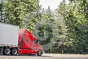 Bright red big rig long haul semi truck with refrigerator semi trailer moving on the road with green trees on the side