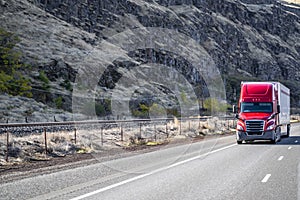 Bright red big rig long hauler semi truck with dry van semi trailer running on the road along the rocky mountain range