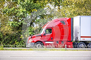 Bright red big rig semi truck with dry van semi trailer running quickly on the highway with green trees on the side for timely