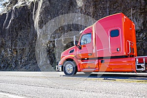 Bright red big rig semi truck with extended cab transporting cargo driving on the mountain road along the rock cliff with