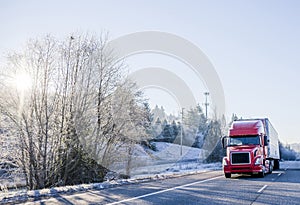 Bright red big rig semi truck with refrigerator semi trailer transporting cargo on straight winter highway frosty hill trees