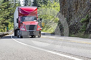 Bright red big rig semi truck with roof spoiler transporting cargo in dry van semi trailer driving on the winding mountain road