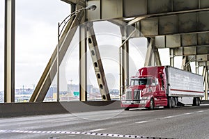 Bright red big rig semi truck transporting cargo in refrigerator semi trailer moving on the two levels bridge across Willamette