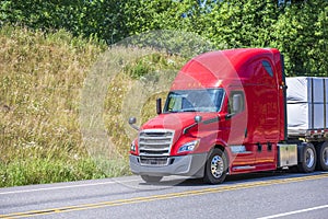 Bright red bonnet big rig semi truck transporting commercial cargo on flat bed semi trailer running on the road with hillside