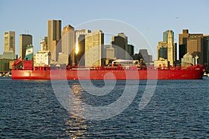 Bright red cargo ship travels in front of Boston Harbor and the Boston skyline at sunrise as seen from South Boston, Massachusetts