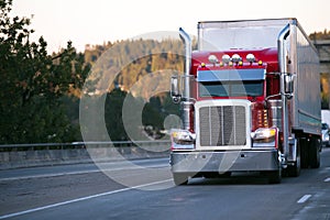 Bright red classic big rig semi truck with trailer move on evening road with turn on headlight