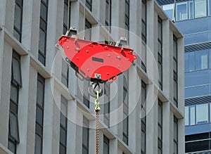 A bright red crane hook and pulley lifting a suspended metal chain on a construction site with urban modern buildings