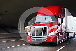 Bright red modern big rig semi truck with dry van trailer running under concrete bridge