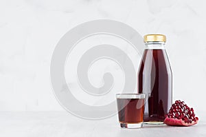 Bright red pomegranate juice in glass bottle mock up with wine glass, fruit grains on white wood table in light interior, template