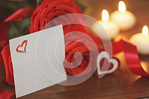 Three bright red roses, sign heart, satin ribbon, sheet of paper and burning candles on wooden background closeup.