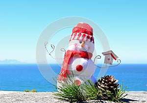 Bright red and white handmade snowman soft toy fleece snowman by bright blue sea horizon background with pine cone & pine needles