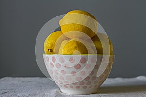 Bright ripe yellow lemons in a white porcelain bowl with pink balls in drops of water on a baggy cloth on a gray background.