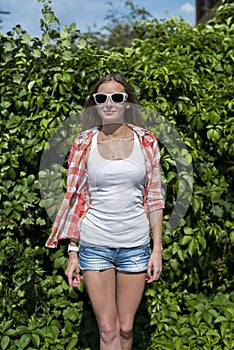 Bright sexy, beautiful young girl, wearing glasses posing in front of a green garden bushes,