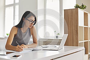 Bright student taking notes in notebook and using laptop computer for e-learning