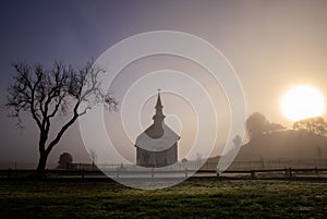 Bright sun and dark fog over small church and tree in rural countryside at dawn photo