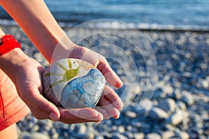 Bright sun painted on pebble in the hands of a child on the background of a pebble beach. Pebbles and sea background