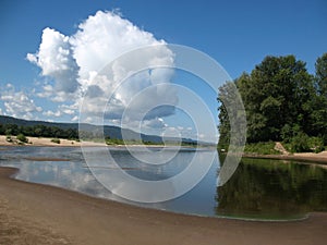 Bright sunny day. Calm river, sandy beach. Blue sky, big cloud