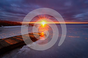 a bright sunset with the sun in a narrow skylight with sunbeams and a dramatic cloudy sky over a frozen lake with an old wooden