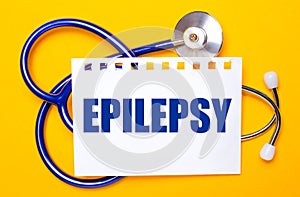 On a bright yellow background, a blue stethoscope and a sheet of paper with the text EPILEPSY. Medical concept