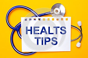 On a bright yellow background, a blue stethoscope and a sheet of paper with the text HEALTS TIPS. Medical concept
