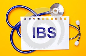 On a bright yellow background, a blue stethoscope and a sheet of paper with the text IBS Irritable Bowel Syndrome. Medical concept