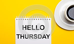 On a bright yellow background, a white cup with coffee and a white notepad with the words HELLO THURSDAY