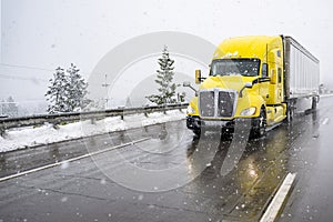 Bright yellow big rig semi-truck transporting cargo in dry van semi trailer driving on the wet slippery highway road with winter