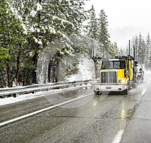 Bright yellow day cab big rig car hauler semi truck with empty semi trailer driving on the winter slippery highway road at snowing