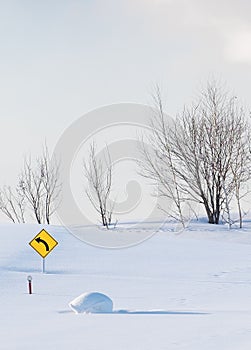 Bright Yellow left turn sign placed and drowned in a snowy field with bare trees on the background.