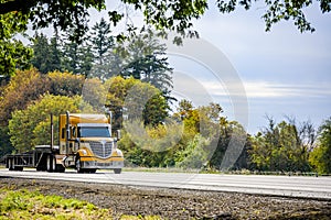 Big rig yellow bonnet semi truck with chrome grille and exhaust pipes transporting step down semi trailer running on the autumn