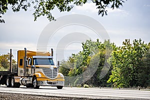 Stylish big rig yellow semi truck with chrome accessories transporting step down semi trailer moving on the road with trees on the